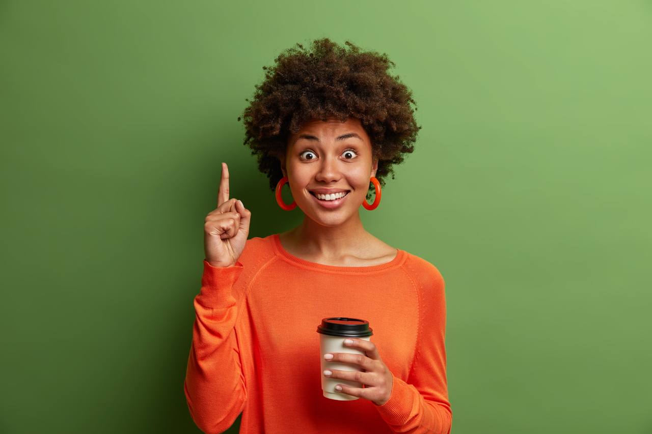 Glad African American woman with curly hair, drinks takeaway coffee, points fore finger above, satisfied to visit cozy cafeteria, has toothy smile, recommends something, poses over green studio wall