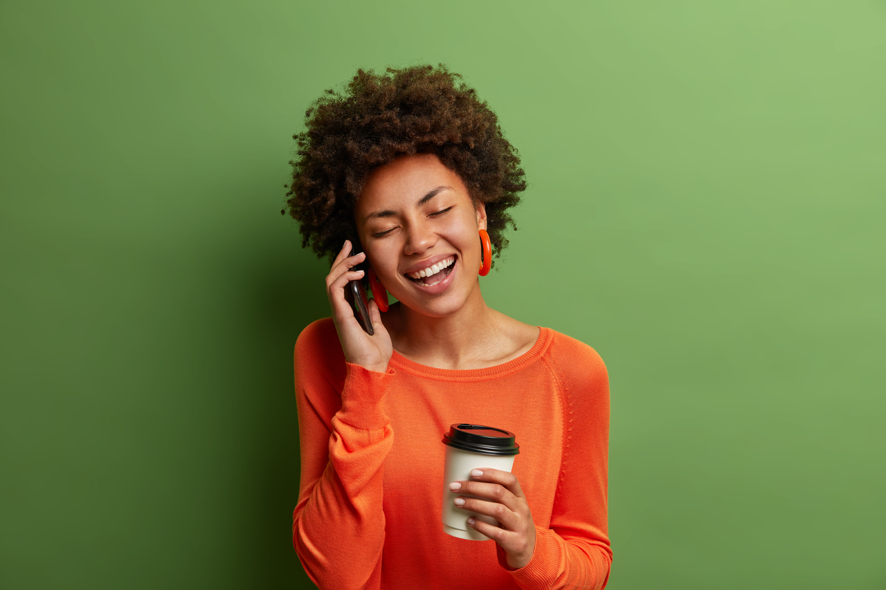 Portrait of funny woman with curly hair, laughs happily, has phone conversation, being amused by friend, drinks coffee from disposable cup, dressed casually, closes eyes, green vivid background